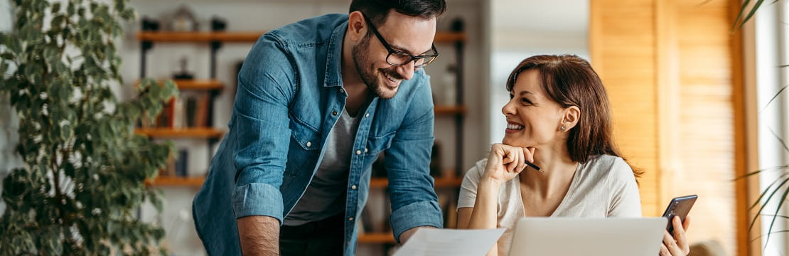A smiling couple reviews a document; there is a laptop before them on a table